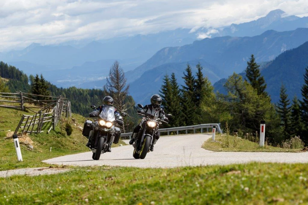 Two motorcyclists riding on a winding mountain road with trees and mountains in the background