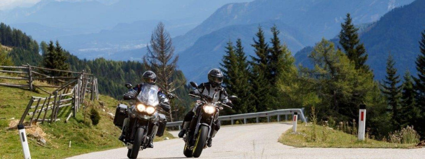 Zwei Motorradfahrer auf einer kurvenreichen Bergstraße mit Bergen und Bäumen im Hintergrund