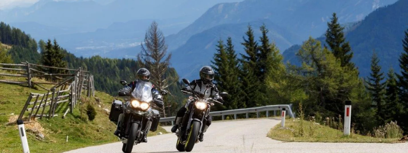 6/7 Nächte - Frühlings-Motorrad-Spass in Val di Sole Twee motorrijders op een kronkelende bergweg met bomen en bergen op de achtergrond
