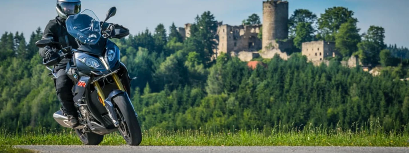 Kurvenreich Motorradwoche 2026/27 Motorrijder op weg met bos en kasteelruïne op de achtergrond