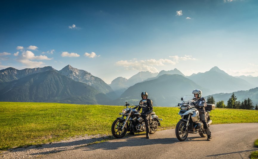 Zwei Motorradfahrer auf Landstraße mit grünen Wiesen und Alpen im Hintergrund
