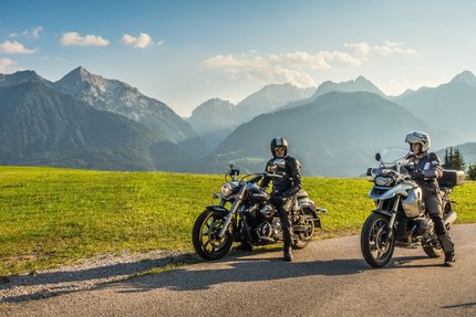 Zwei Motorradfahrer auf Landstraße mit grünen Wiesen und Alpen im Hintergrund