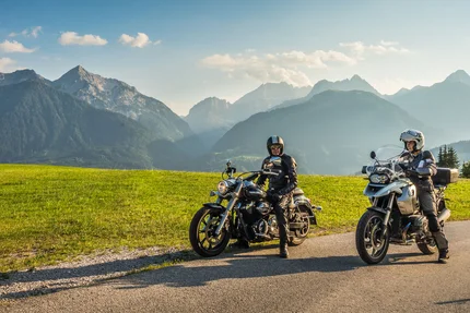 Twee motorrijders op een landweg met groene velden en Alpen op de achtergrond