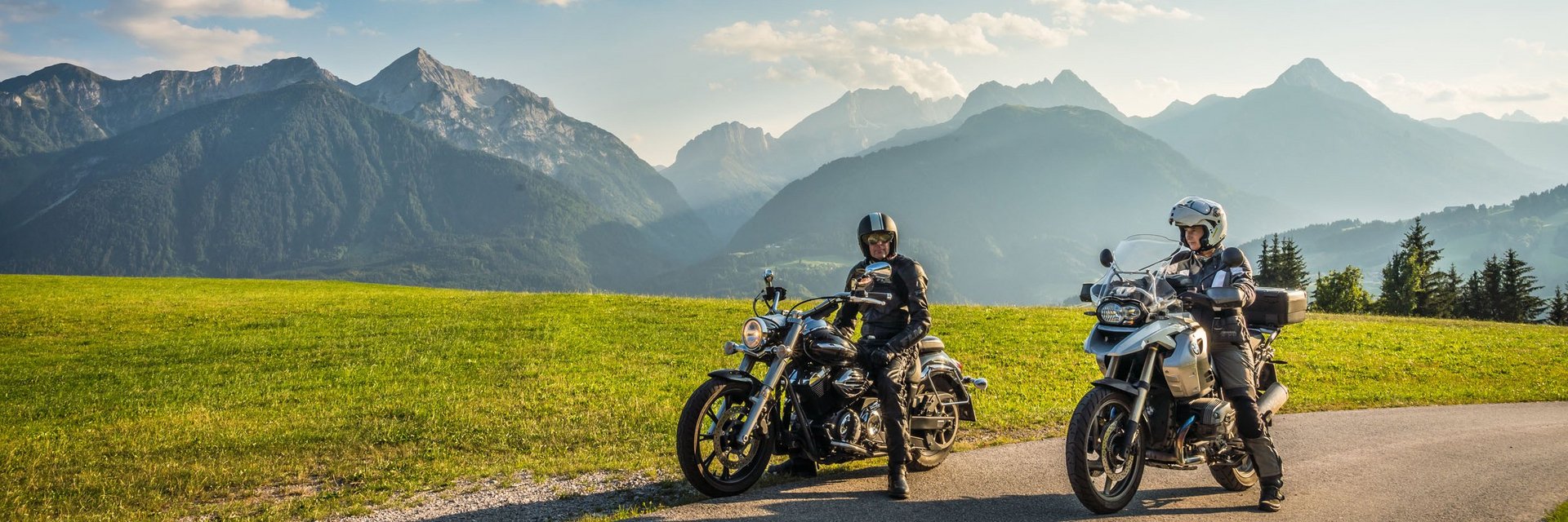 Zwei Motorradfahrer auf Landstraße mit grünen Wiesen und Alpen im Hintergrund