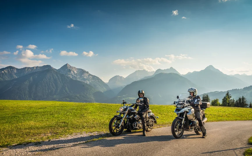 Two motorcyclists on rural road with green fields and Alps in the background