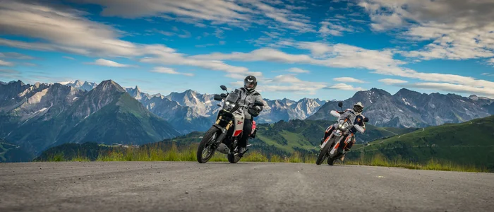 Two motorcyclists on mountain road with Alps in background under cloudy sky