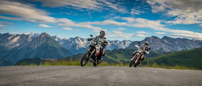 Zwei Motorradfahrer auf Bergstraße mit Alpen im Hintergrund bei bewölktem Himmel