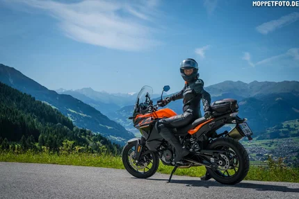 Motorcyclist in black gear on orange bike with mountains in background