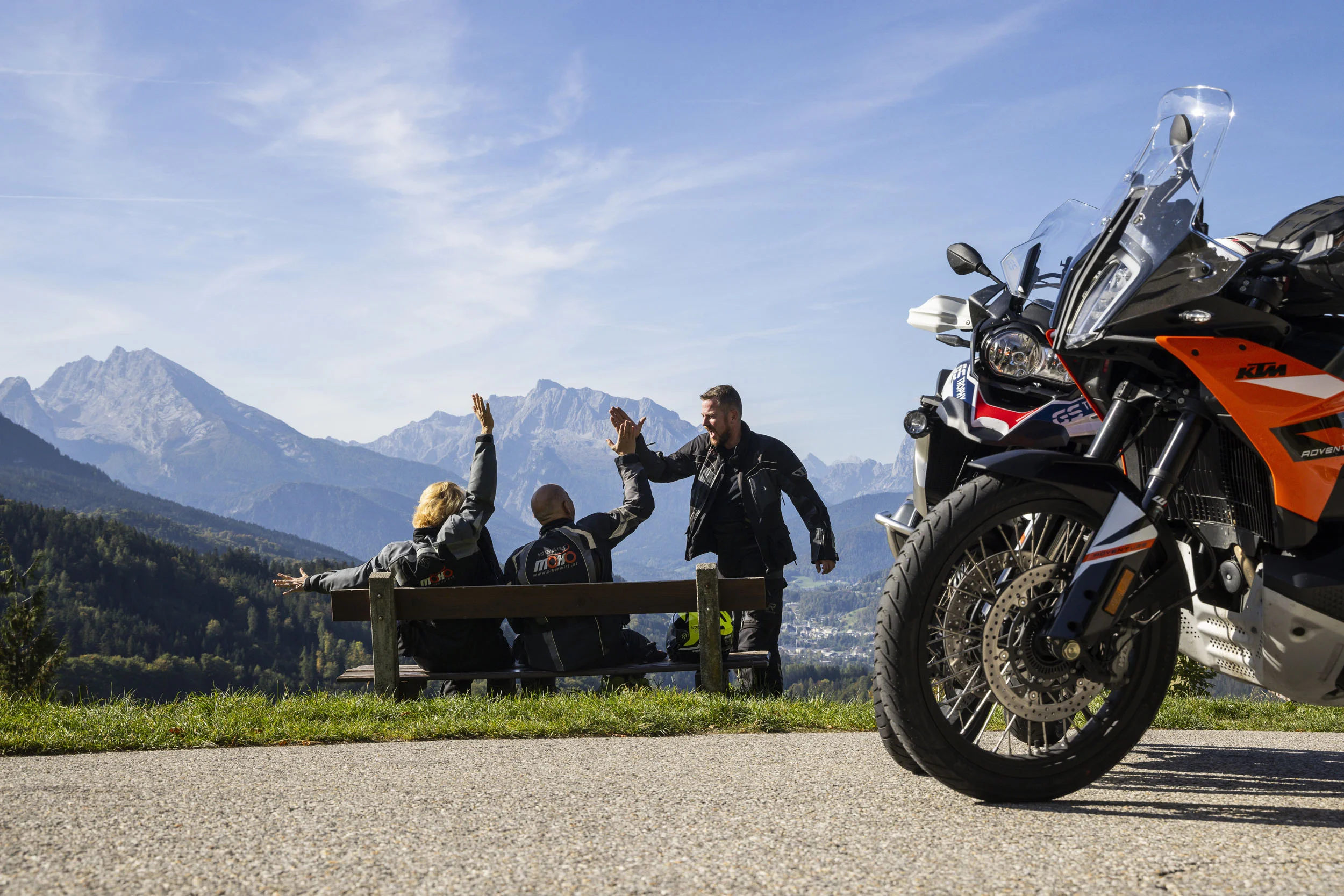 My MoHo-App © Heiko Mandl Drei Motorradfahrer geben sich vor Berglandschaft ein Highfive auf einer Bank