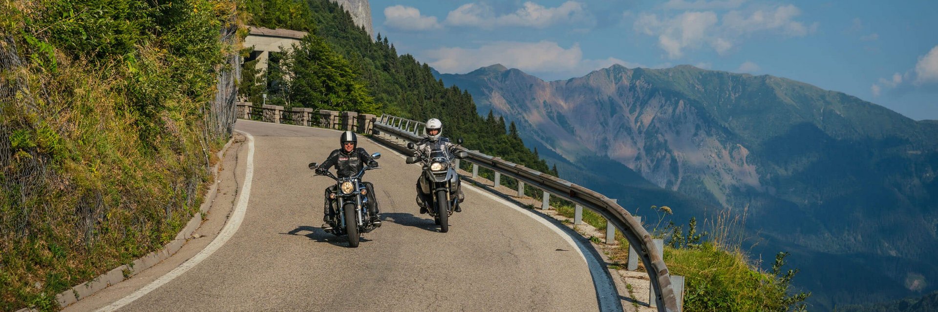 Zwei Motorradfahrer auf kurviger Bergstraße mit Bergen im Hintergrund.