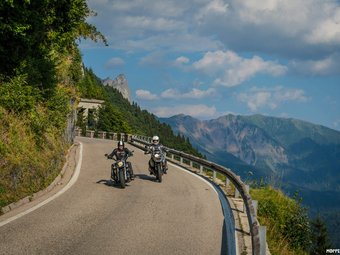 Zwei Motorradfahrer auf kurviger Bergstraße mit Bergen im Hintergrund.