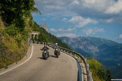 Zwei Motorradfahrer auf kurviger Bergstraße mit Bergen im Hintergrund.