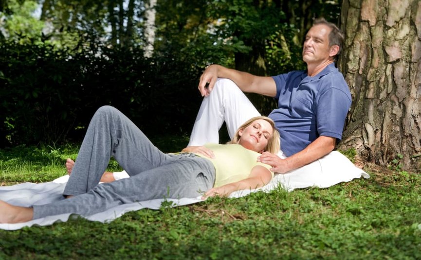 Mann lehnt an Baum, Frau liegt mit geschlossenen Augen auf seinem Schoß im Park