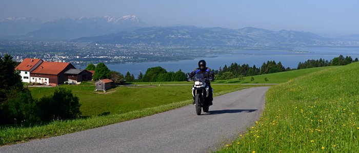 Motorradfahrer auf Landstraße mit Alpenpanorama und See im Hintergrund