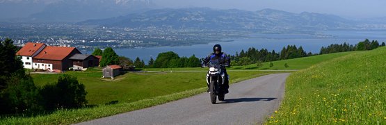 Motorradfahrer auf Landstraße mit Alpenpanorama und See im Hintergrund