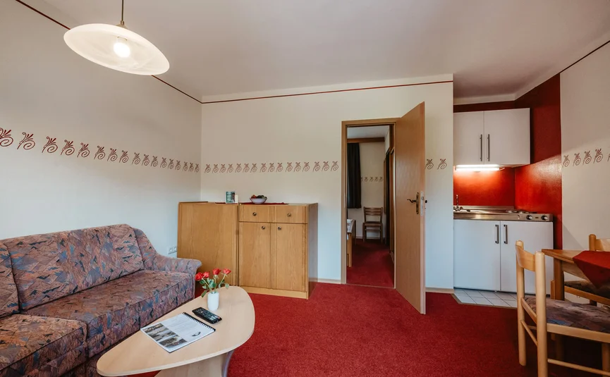 Living room with sofa, view into bedroom and small kitchen in red-beige design