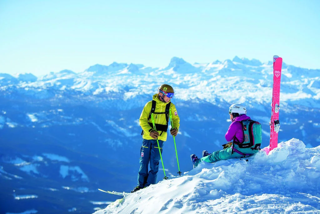 Two skiers in colorful gear on snowy mountain with scenic mountain backdrop