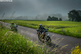 Zwei Motorradfahrer auf Landstraße bei nebligem Wetter und grüner Landschaft
