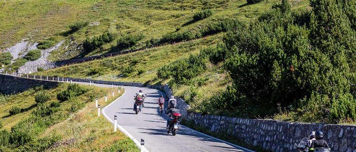 Motorrijders op bergweg met groene hellingen en graffiti op een muur