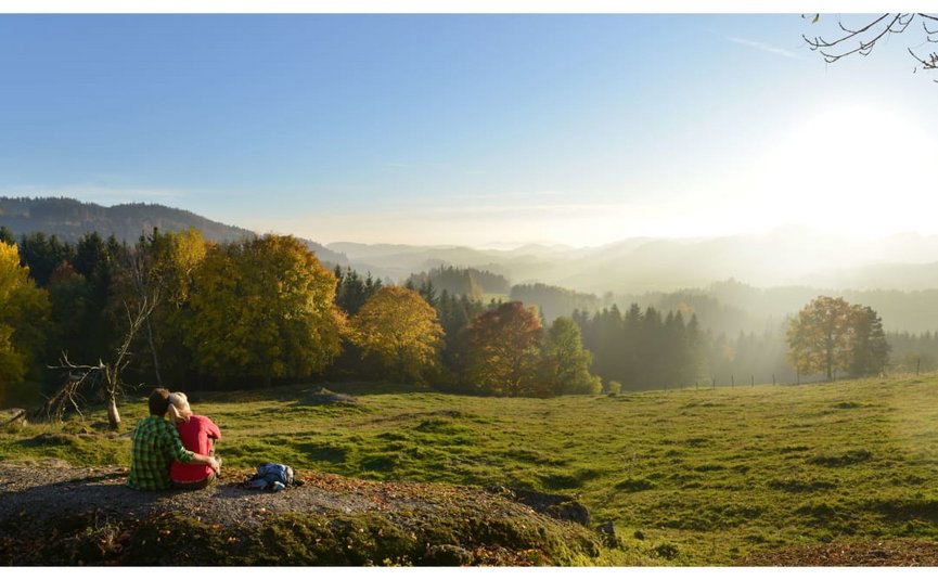 Kraftquelle Naturparadies Paar sitzt auf einem Felsen und blickt auf sonnige Herbstlandschaft mit Wald und Bergen