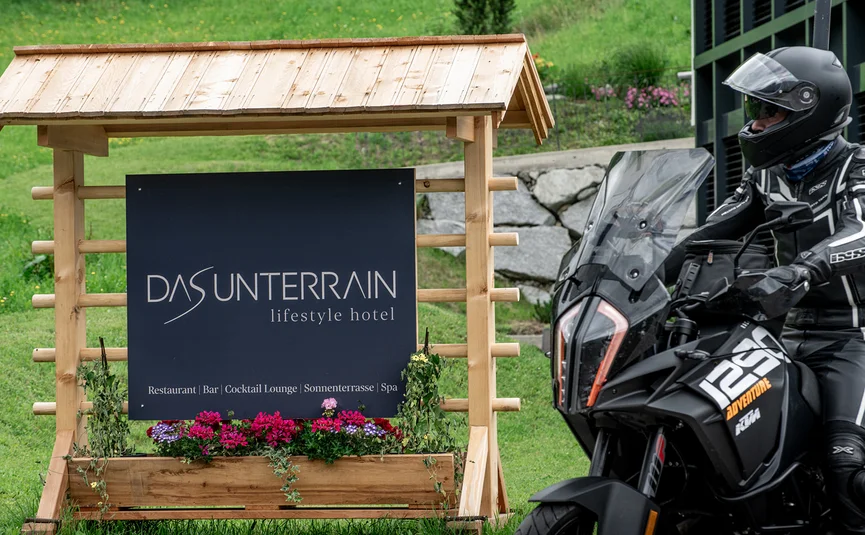 Motorradfahrer neben Holzschild vom Hotel Das Unterrain mit Blumen