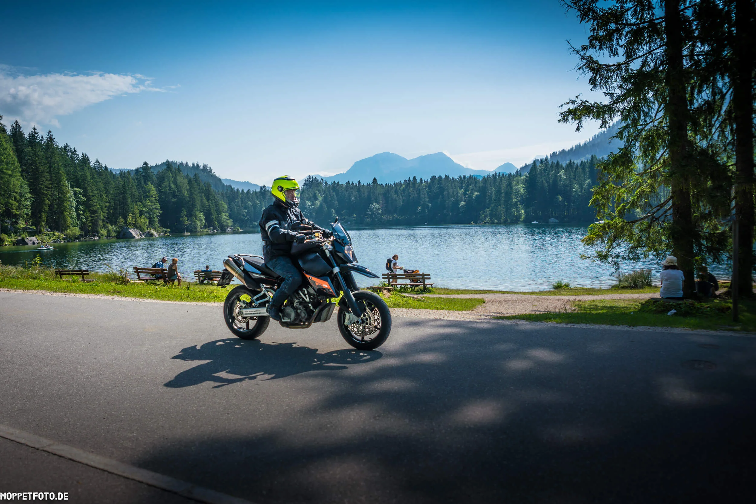 Motorradfahrer mit gelbem Helm fährt an einem See mit Bergen und Bäumen vorbei