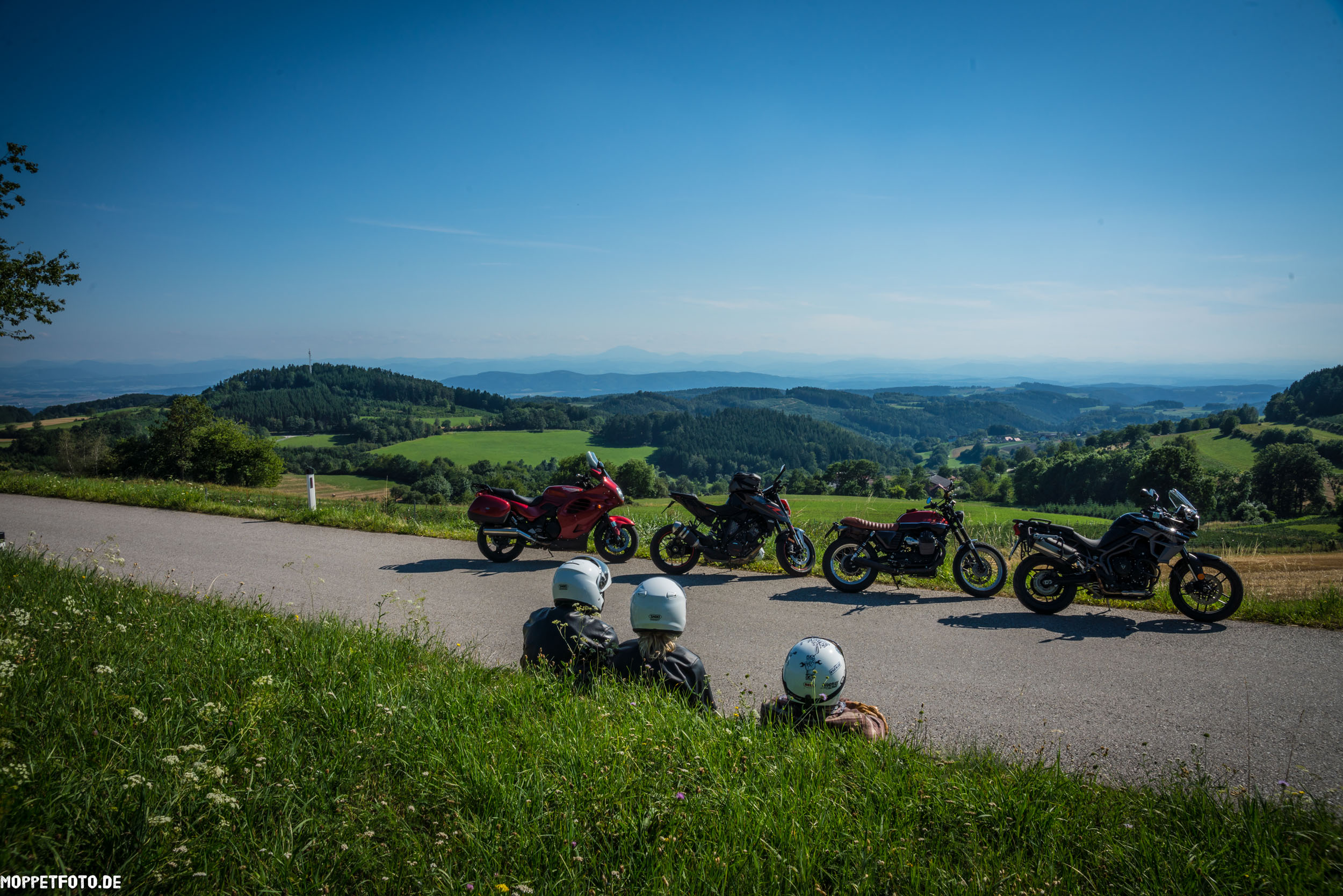 Drei Motorradfahrer sitzen am Straßenrand mit Blick auf grüne Hügel und blaue Berge