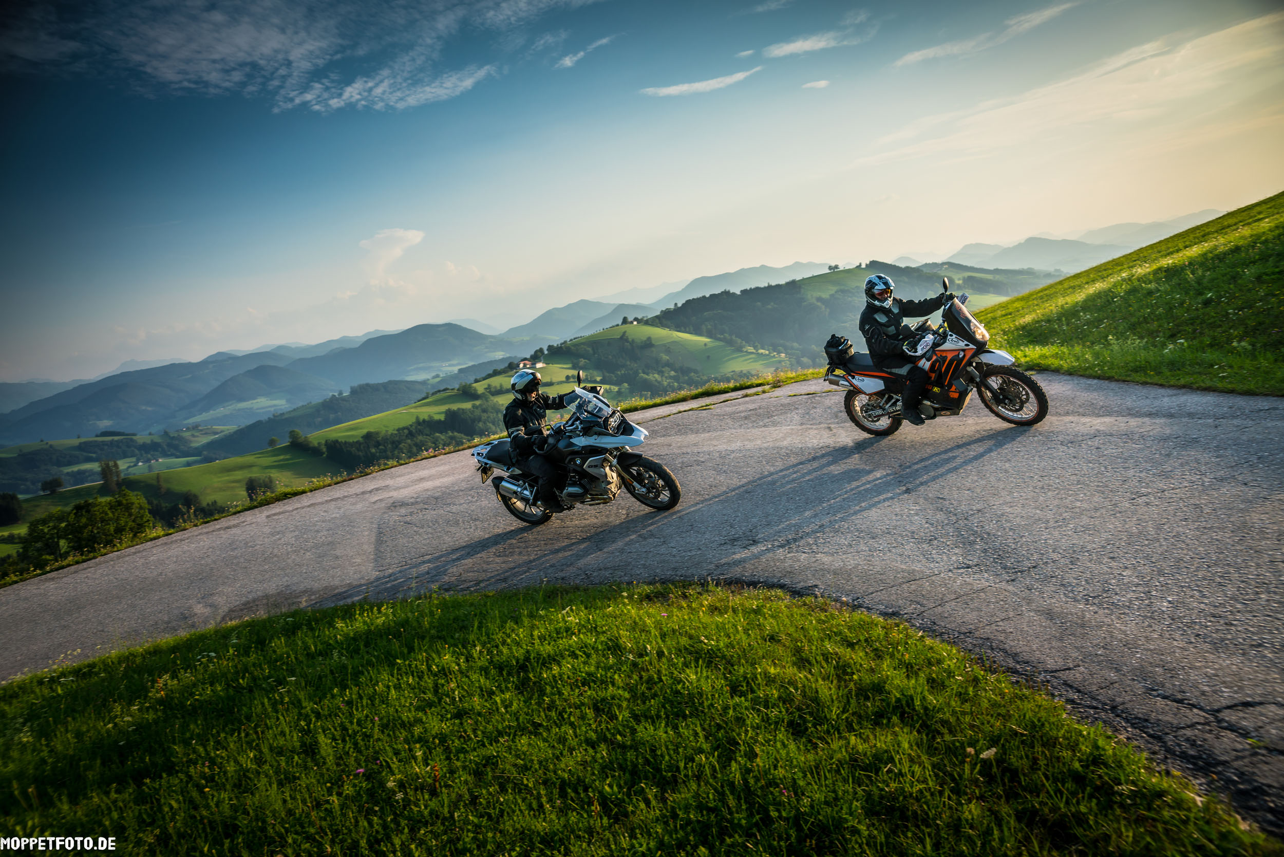 Jouw motorervaring © Moppetfoto.de Twee motorrijders op een kronkelende bergweg met groene heuvels en blauwe lucht