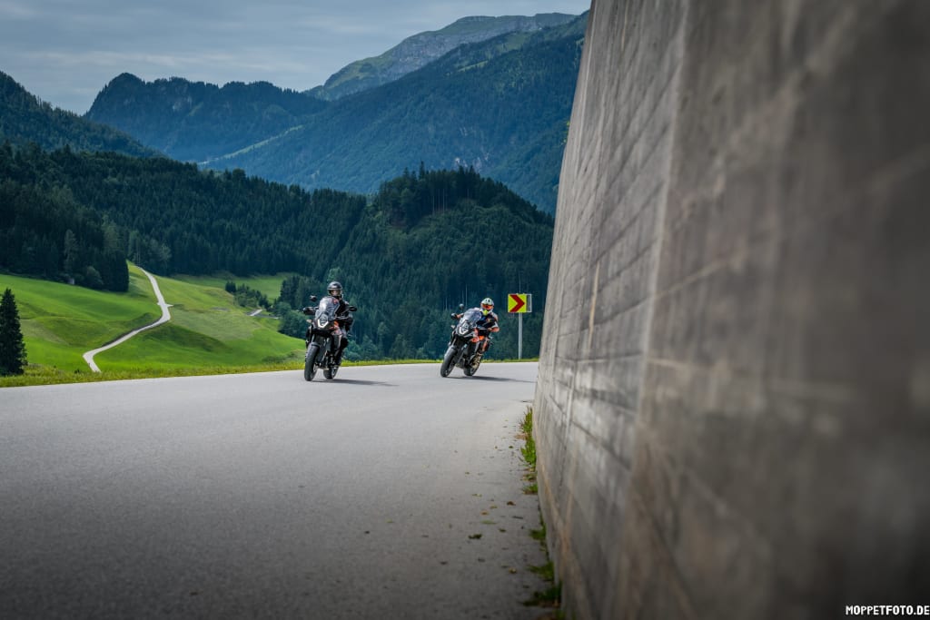 Zwei Motorradfahrer auf kurviger Bergstraße mit bewaldeten Bergen im Hintergrund