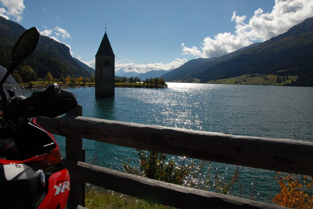 Kirchturm ragt aus einem See in den Alpen mit Motorrad im Vordergrund