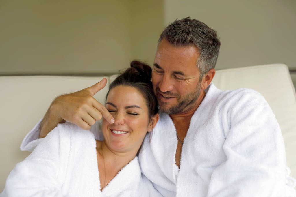 Paar in weißen Bademänteln lacht, Mann berührt liebevoll die Nase der Frau