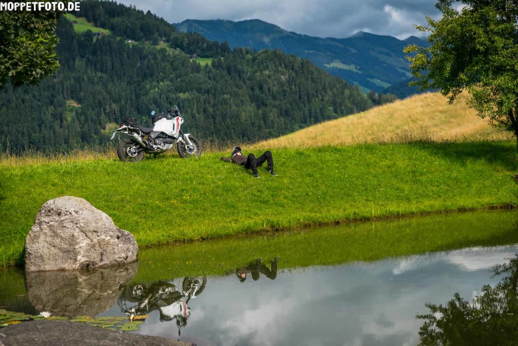 Motorrad und Person liegen auf Gras neben einem See und Bergen im Hintergrund
