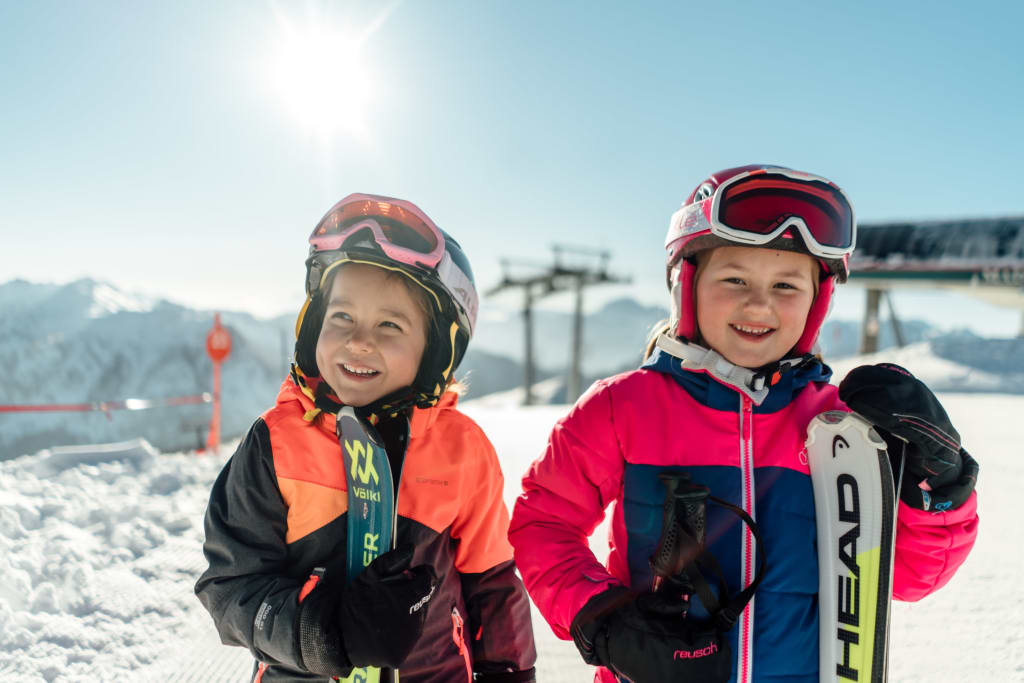 Zwei lachende Kinder mit Skihelmen und Skiern im Schnee bei Sonnenschein