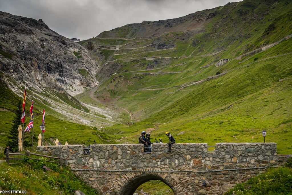 Steinbrücke mit Motorradfahrern vor grünen Bergen und Serpentinenstraße