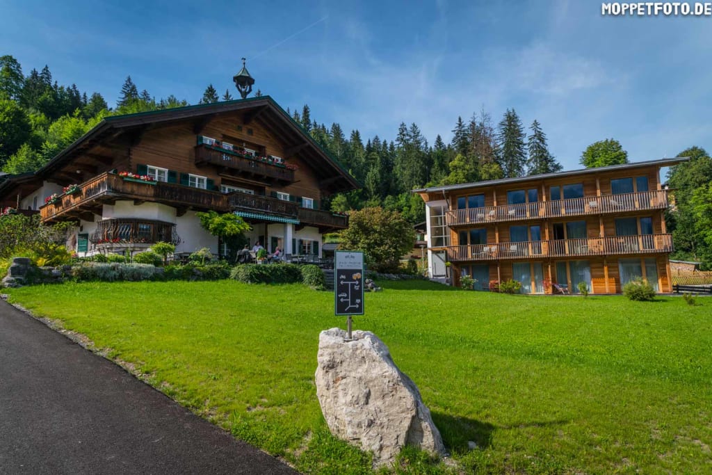 Traditionelles Holzhaus und modernes Gebäude auf grünem Hügel vor Wald