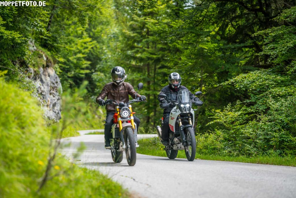 Zwei Motorradfahrer fahren auf kurvenreicher Straße durch grünen Wald