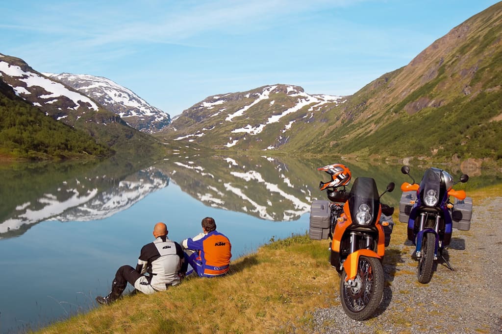 Twee motorrijders zitten aan een meer met besneeuwde bergen en weerspiegelde lucht