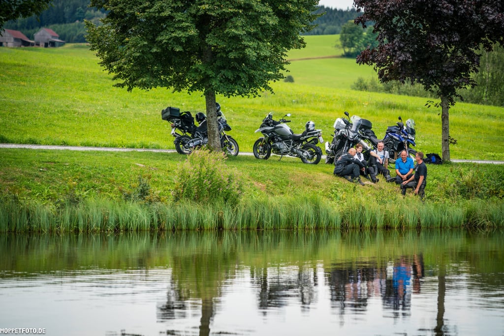 Fünf Menschen sitzen am Seeufer neben Motorrädern auf einer grünen Wiese