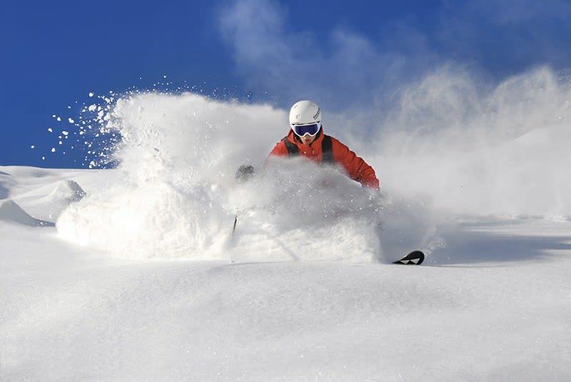 Skifahrer in roter Jacke fährt durch tiefen Pulverschnee bei klarem Himmel
