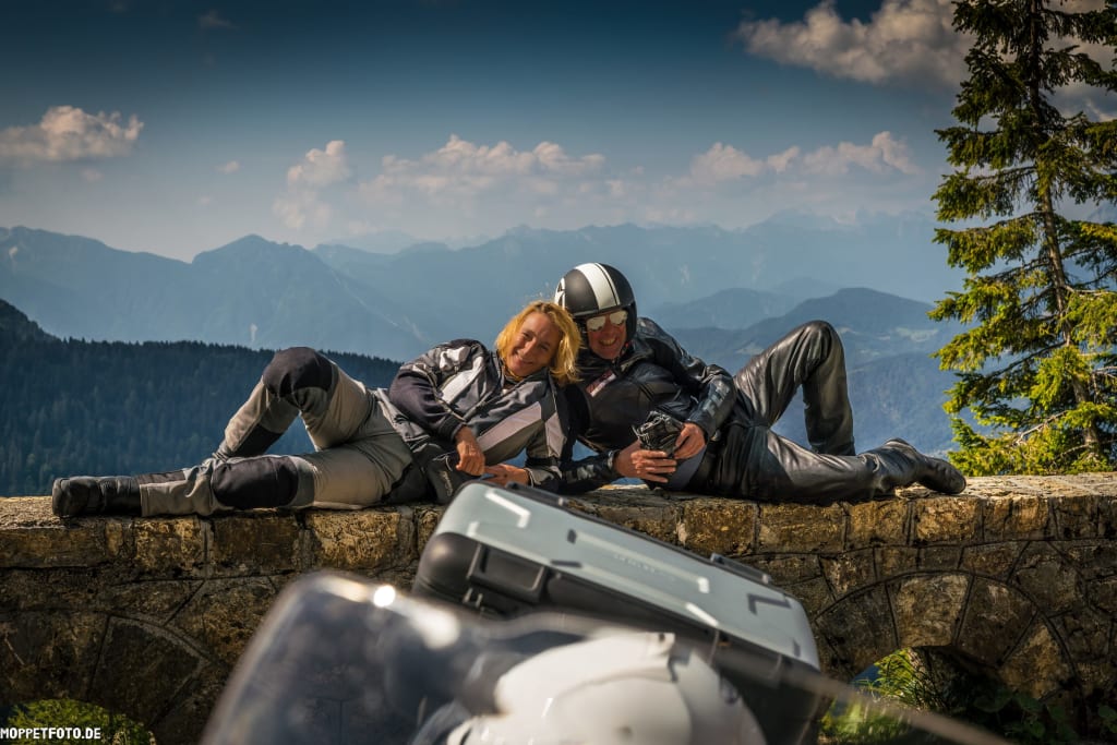 Zwei Motorradfahrer in Schutzkleidung liegen auf einer Steinmauer mit Bergpanorama