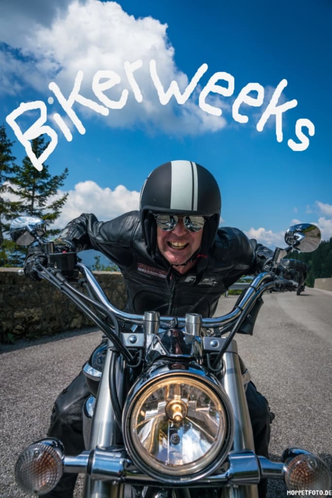 Lächelnder Motorradfahrer in Lederjacke mit Helm vor blauem Himmel mit Bikerweeks-Schrift