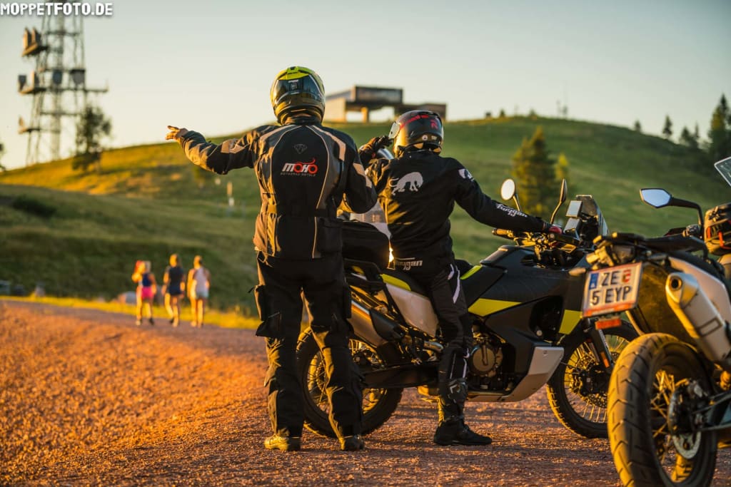 Zwei Motorradfahrer in Schutzkleidung zeigen auf einen Hügel bei Sonnenuntergang