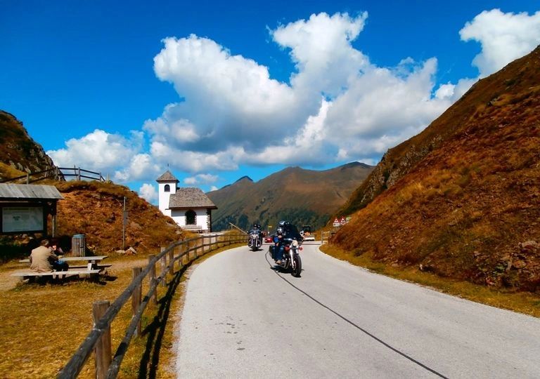 Motorcyclists riding on mountain road beside chapel under sunny sky